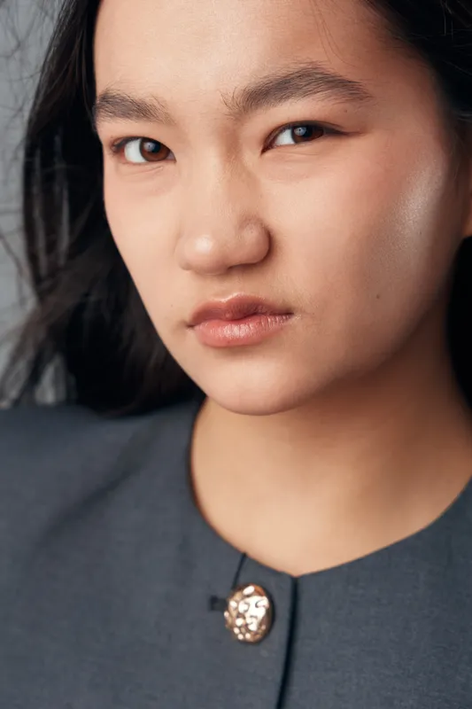 Portrait of serious woman with black hair and natural makeup looking at camera in studio, wearing gray shirt with decorative button on collar. Closeup of face and expression.