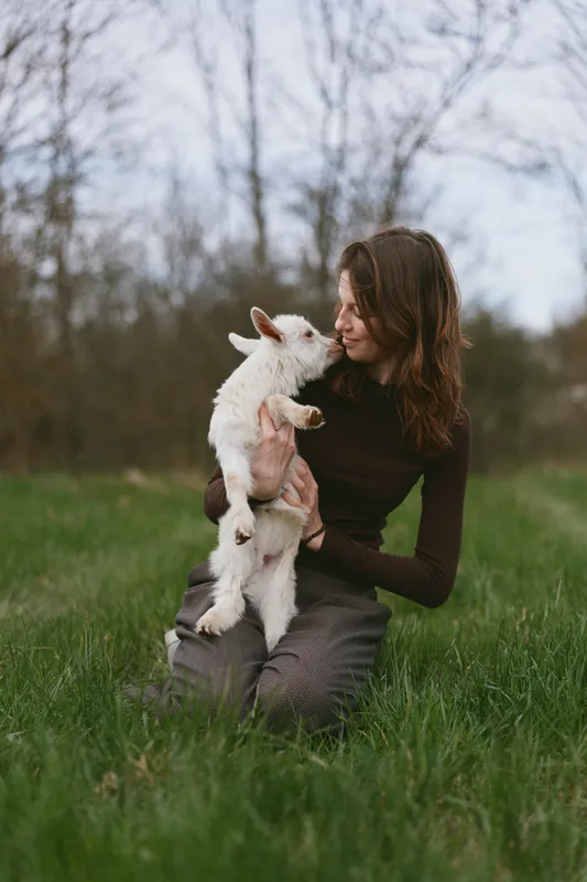 A young woman with long brown hair kneels in the grass holding a small white baby goat while looking at it affectionately in a natural outdoor setting during early spring.