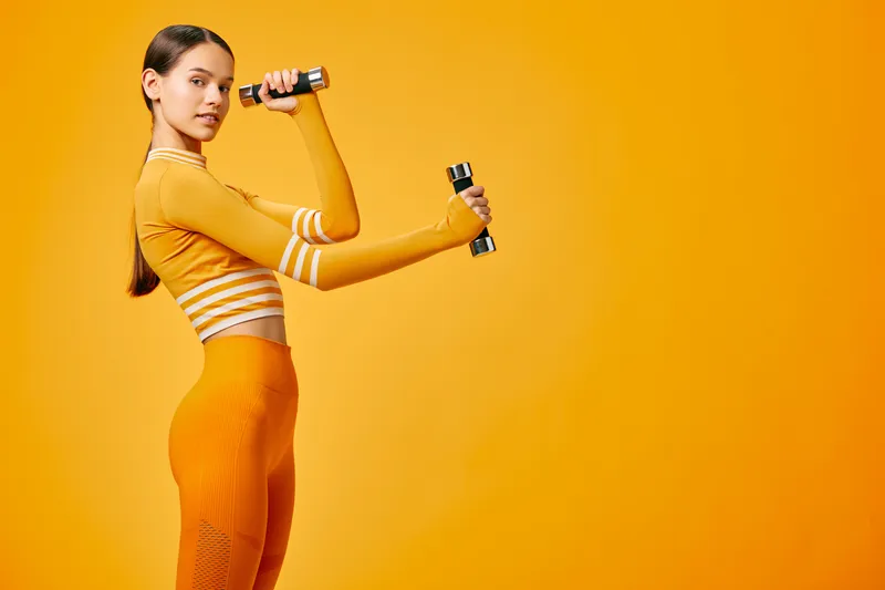 Fit young woman exercising with small dumbbells in a monochrome orange studio, wearing matching activewear, poised in a strength pose, theme woman fitness, sport.