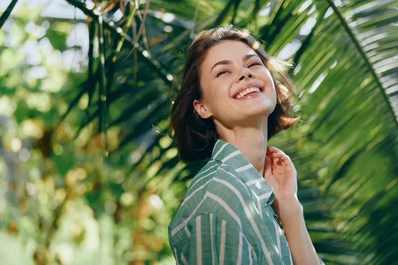 Portrait of a smiling woman standing among tropical leaves, sunlight on her face, exuding relaxed natural charm and outdoor warmth.