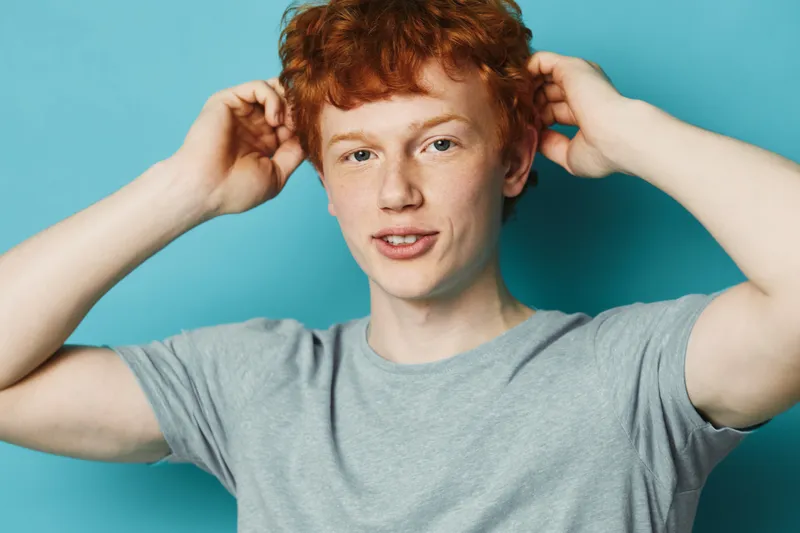 Young man with red hair and freckles posing with hands behind head against blue background, casual style and confident expression indoors with simple gray t shirt.