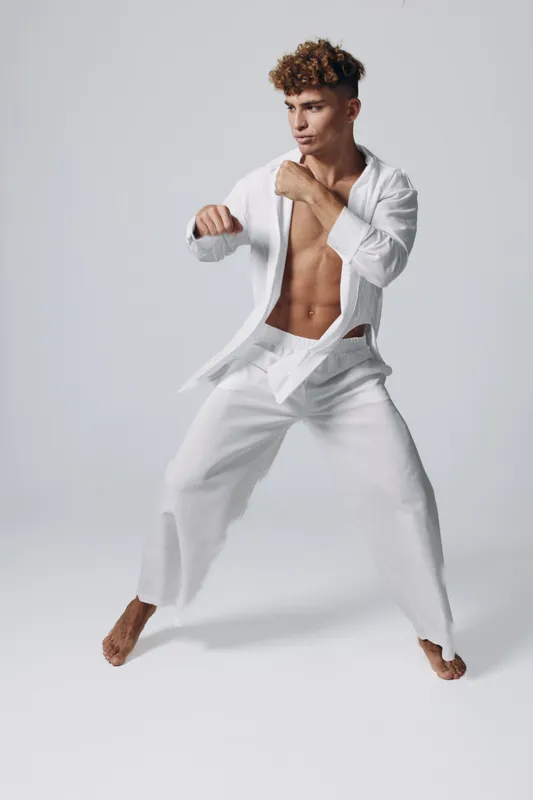 Young man practicing martial arts in white gi with bare chest and focused expression, demonstrating fighting stance and punches on plain background.
