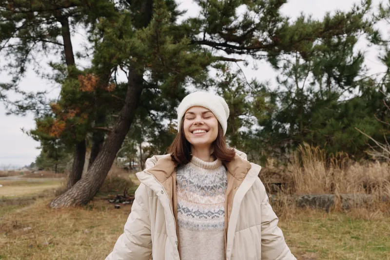 Woman outdoor winter smiling coat scenery as she wears a cozy knit sweater and beanie, standing in a field with trees, capturing a bright, calm moment in fresh air