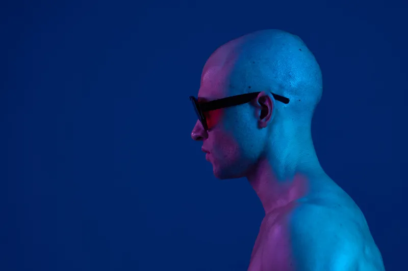 A bald man with sunglasses poses strikingly in a cool blue tone, exuding a confident and modern vibe with energetic lighting effects.