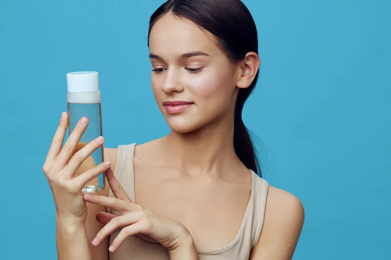 Young woman examining a skincare bottle against a blue background, showcasing a fresh and healthy look, perfect for beauty and wellness promotions