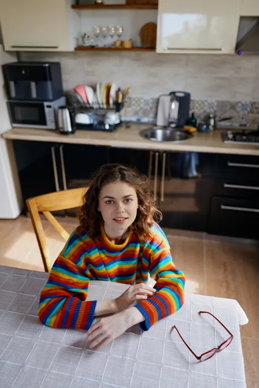 Young woman with curly hair sits at a kitchen table wearing a colorful striped sweater, studying a notebook with a pair of glasses nearby in a bright home scene