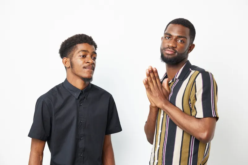 Two black men expressing different emotions, one with a thoughtful smile and the other looking contemplative, against a simple, light background.