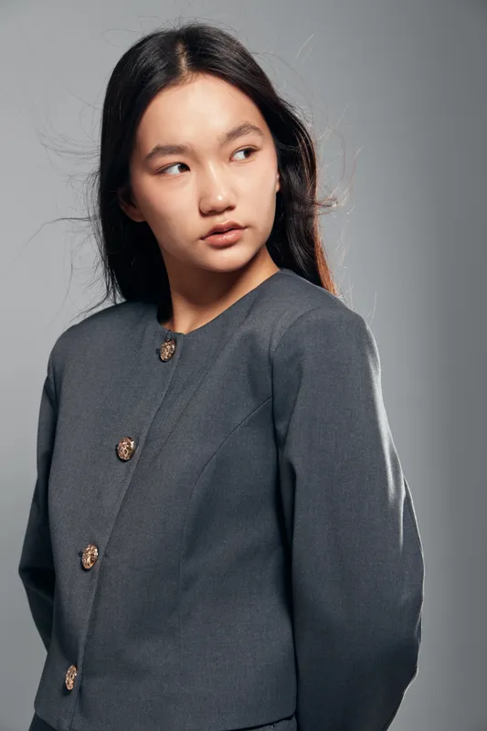 Young Asian woman in gray jacket with decorative buttons looking sideways with neutral expression standing in studio with plain background and soft lighting.