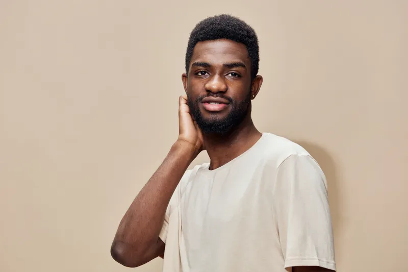 Young African man in a light t shirt, smiling and touching his neck, with a neutral beige background, illustrating confidence and modern style