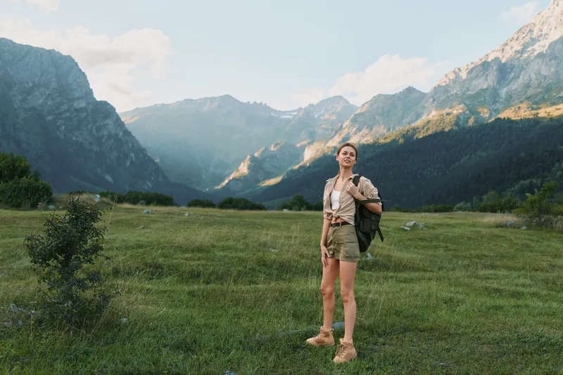 Cheerful woman outdoors, hiking in a valley with mountains, sunlit meadow, casual attire, backpack, relaxed stance, natural colors, adventurous mood