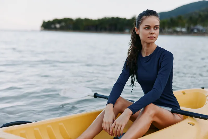 A young woman sits in a yellow rowboat on calm water, wearing a navy long sleeve top, with wind swept hair and a thoughtful gaze against a distant shore.