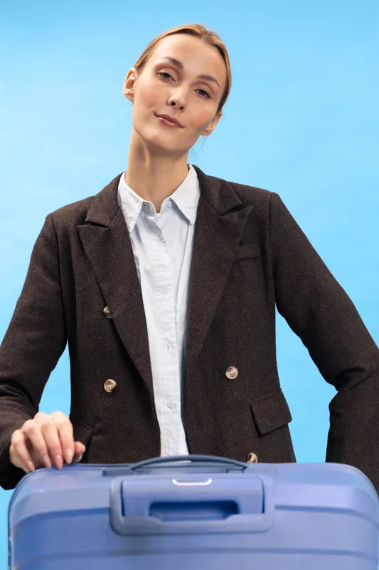 Confident businesswoman in dark blazer stands behind blue suitcase on light blue background. Concept of travel, business trip, and professional lifestyle with serious expression.