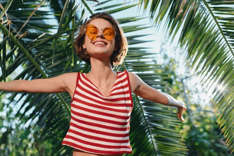 A joyful young woman stands outdoors amid tropical palms, wearing a red striped crop top and round sunglasses. She smiles brightly, arms outstretched, embodying carefree sunshine.