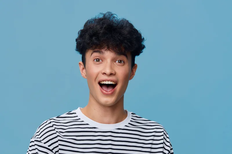 Excited man with curly hair wearing striped shirt smiles brightly against solid blue background, showing enthusiasm and joy in casual style portrait.