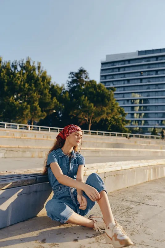 young woman sitting outdoors on concrete steps wearing casual blue denim, red bandana, relaxed pose under sunny clear sky with urban background