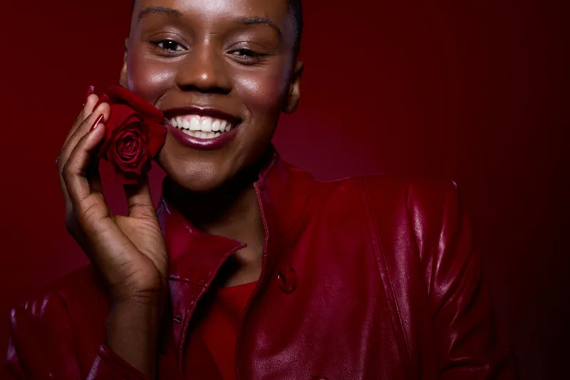 Happy African woman with dark skin smiles holding red rose near face. She wears red leather jacket and red lipstick. Studio shot on red background. Concept of beauty, joy, and elegance.