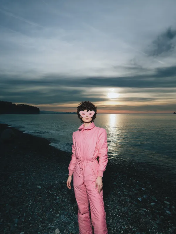 Person in pink jumpsuit stands on a rocky beach at sunset, calm sea and dramatic sky, fashion portrait with moody lighting and distant shoreline.