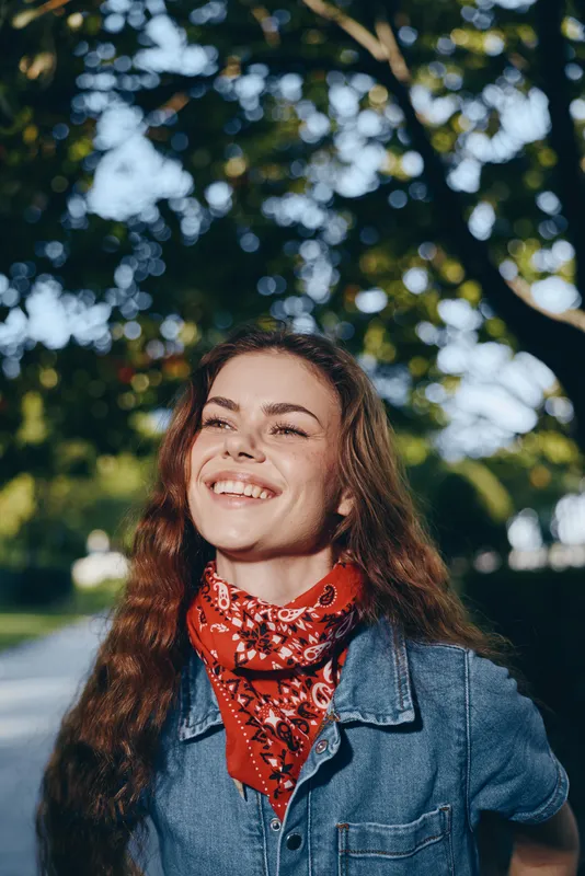 Lifestyle woman smiling in modern boho-western denim street style with a red bandana, film color tones, natural light, and outdoor greenery background.