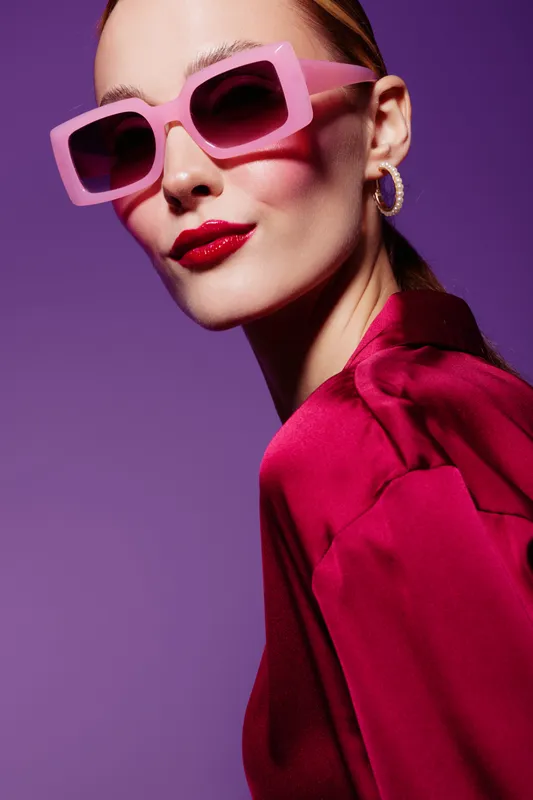 Stylish woman wearing pink sunglasses and red lipstick poses confidently against purple background. Fashion portrait with bold colors and modern accessories. Concept of glamour and style.