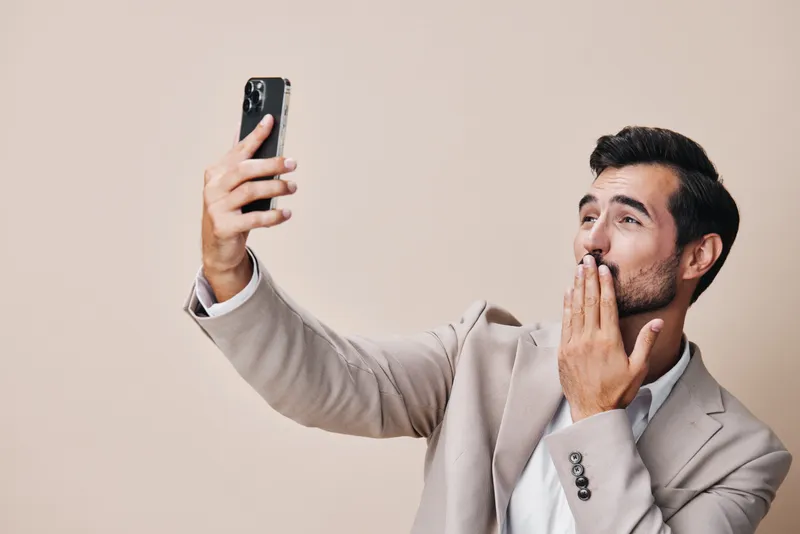 Бородатый мужчина в деловом костюме держит смартфон и улыбается, стоя в студии на сером фоне