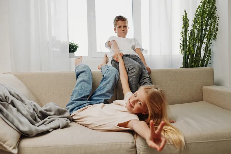 Two playful children having fun on beige couch in bright living room, boy sitting and girl lying down showing peace sign. Concept of childhood joy, sibling play, and home comfort.
