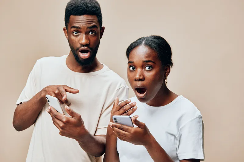 Surprised young couple looking at smartphones in casual clothing, expressing shock and excitement against a neutral background