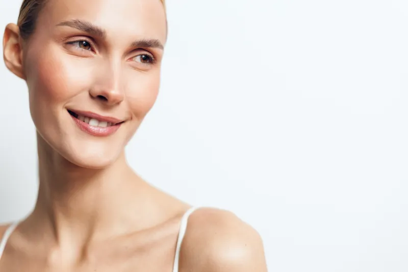 Smiling woman with clear skin looking sideways on a white background. Close-up portrait with natural makeup and soft lighting. Concept of beauty, confidence, and wellness.