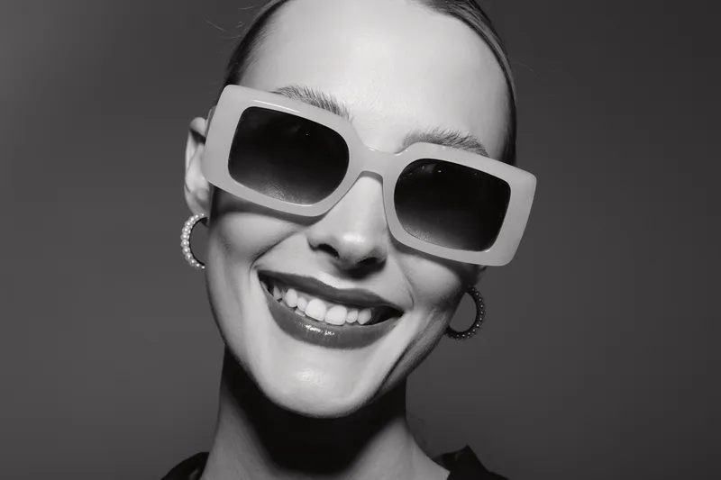 Smiling white woman with fair skin wearing large sunglasses and hoop earrings. Close-up portrait with slicked-back hair in black and white studio lighting.
