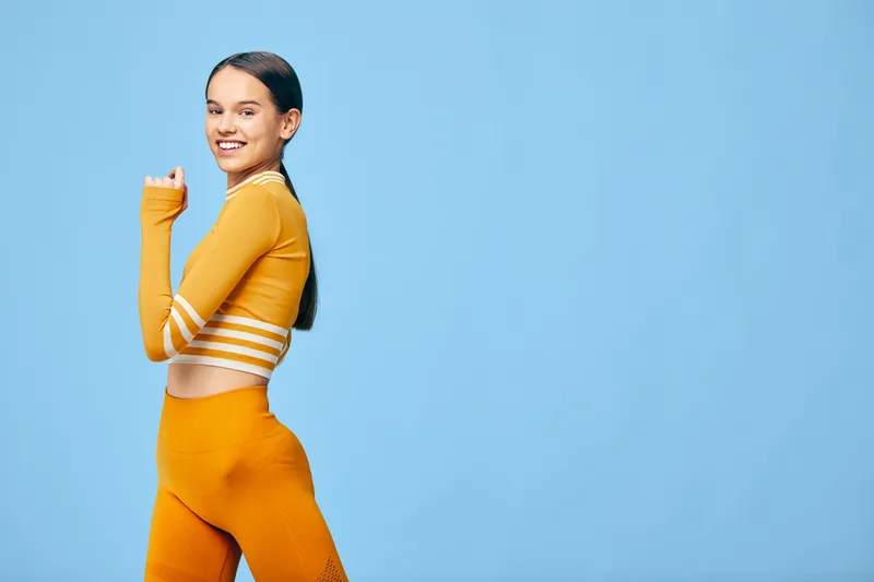 Young woman in orange fitness outfit posing confidently in studio, smiling with ponytail, textured activewear, athletic stance conveying health, empowerment, sport and modern workout lifestyle.