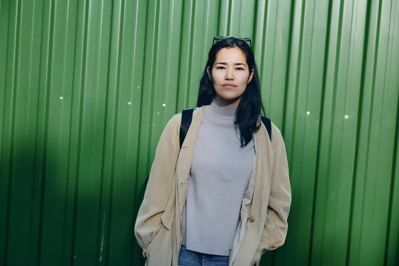 Woman in a cozy sweater and beige jacket stands outdoors against a green wall, showcasing casual fashion and confident style, natural light, minimal background, streetwear vibe, everyday attire,