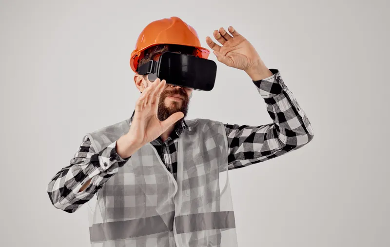Мужчина в оранжевой каске и клетчатой рубашке держит в руках 3D-очки виртуальной реальности, символизируя интернет-технологии в строительстве
