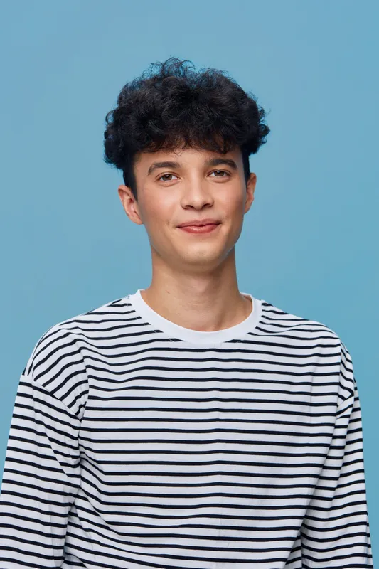 young man with curly hair wearing striped shirt smiling gently against light blue background, casual portrait expressing calm and friendliness
