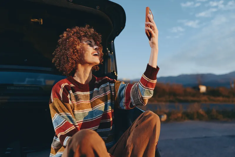 Woman lifestyle, curly hair woman sits by open car outdoors on roadside, smiling and lifting hand toward sunset, casual sweater, roadtrip scenery, evening calm