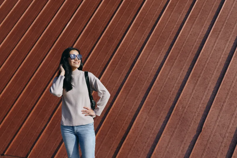 Woman traveler in casual outfit with backpack stands before a rust diagonal wall, sunglasses on, exploring urban scenery, contemporary style, travel mood, street photography.