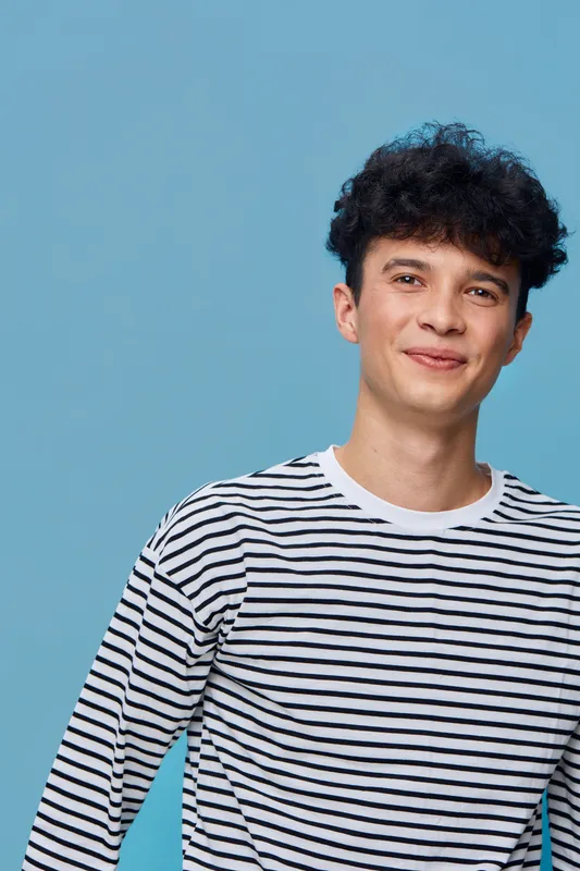 smiling man wearing striped shirt stands against blue background with curly hair and relaxed expression, young adult portrait in casual style