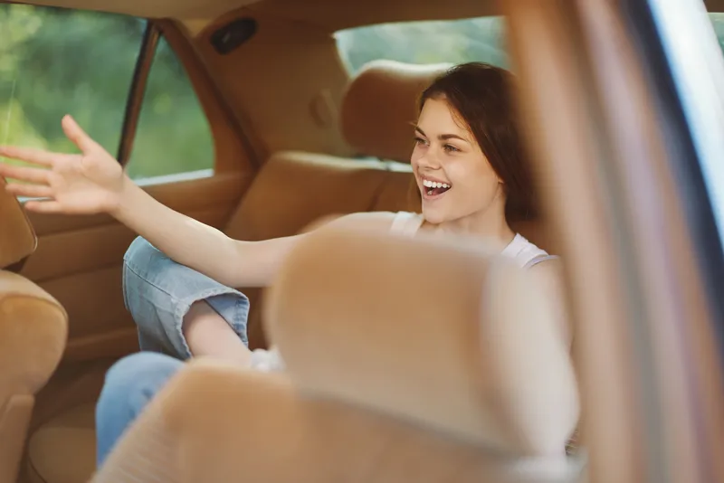 Happy young woman enjoying a road trip, smiling as she talks from the back seat of a car surrounded by warm brown tones and natural outdoor light.
