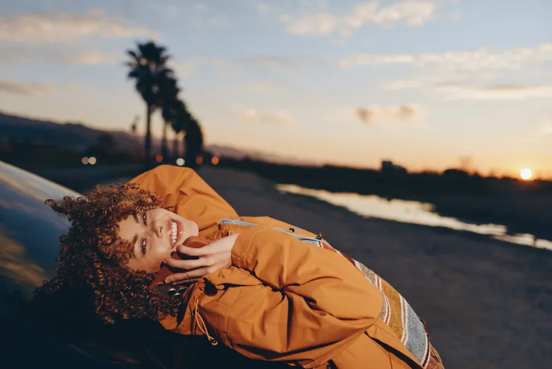 Woman beach lifestyle at sunset, curly hair and yellow jacket, lying on sand by ocean waves, relaxed smile, warm golden sunlight, calm seaside travel mood, breeze, horizon, evening