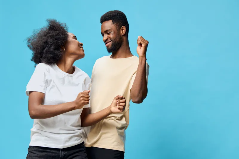 Young couple joyfully dancing together in stylish casual outfits against a vibrant blue background, showcasing happiness and connection