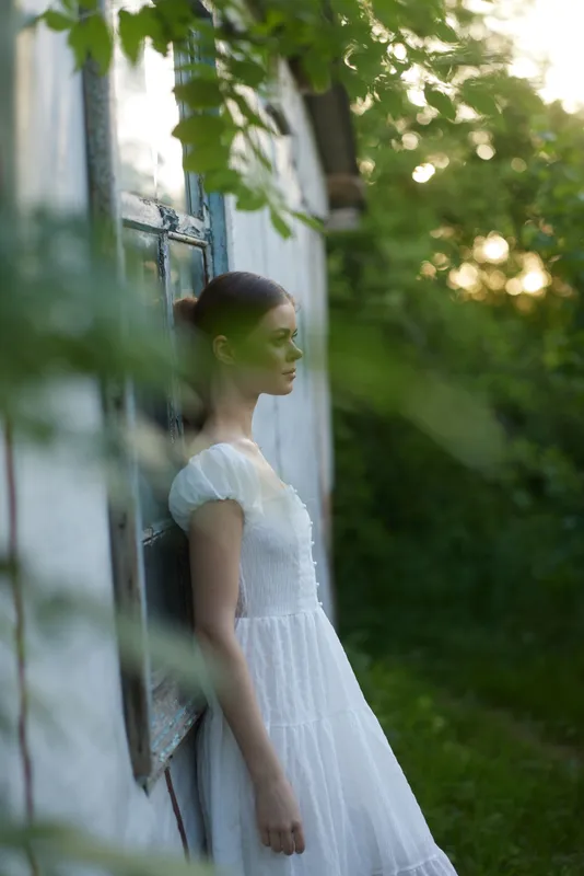 Young woman in a white dress standing by a rustic building, surrounded by greenery, exuding calmness and serenity during sunset