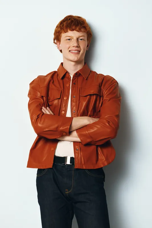 Young man with crossed arms standing against white wall wearing brown leather jacket and casual jeans. Confident, smiling, relaxed posture with stylish outfit and light background.