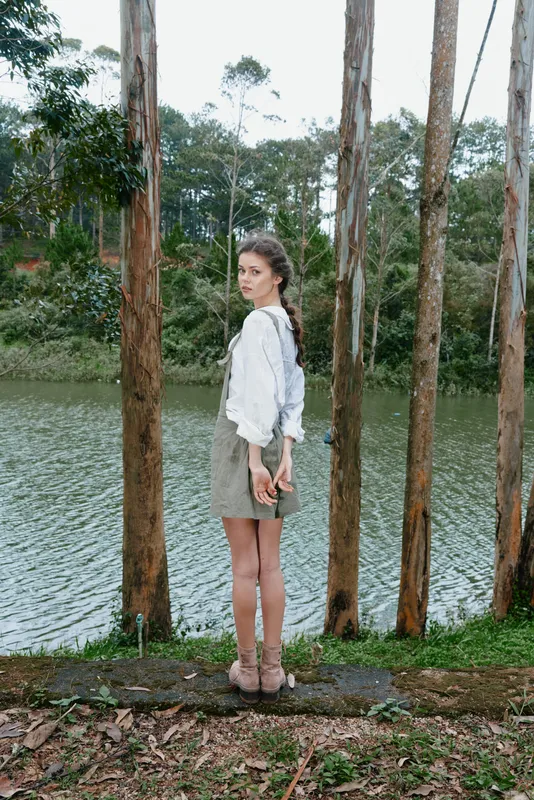 Young woman in a forest standing by a serene lake wearing a light blouse and skirt, showcasing a calm and peaceful demeanor influenced by nature