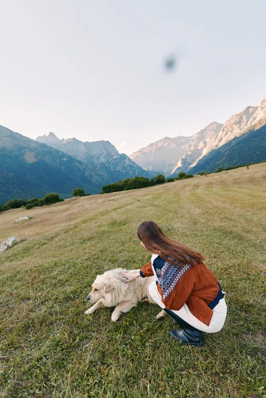 Woman petting white dog in a grassy mountain meadow, outdoor nature scene with landscape views and rolling hills. Relaxed pet owner enjoying fresh air and scenic alpine setting.