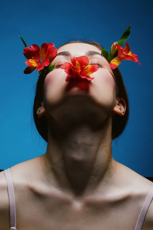 A young woman with an ethereal expression holds vibrant flowers near her face, emphasizing beauty and nature, against a striking blue background