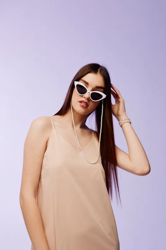 Young woman beauty fashion model wearing trendy white sunglasses and beige dress poses confidently on a light purple background. Concept for style and elegance.