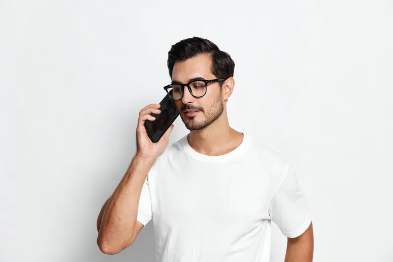Casual young man in white t-shirt wearing glasses talking on smartphone, looking thoughtful with a neutral expression, isolated on plain white background. Modern lifestyle concept.