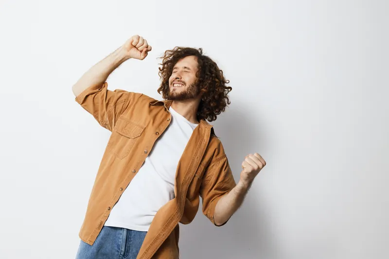 A joyful young man with curly hair celebrating success in a casual outfit, showcasing happiness and positivity against a light background