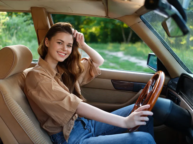 Happy young woman enjoying a road trip in a vintage car, wearing a casual brown shirt and denim jeans, smiling brightly against a lush green background.