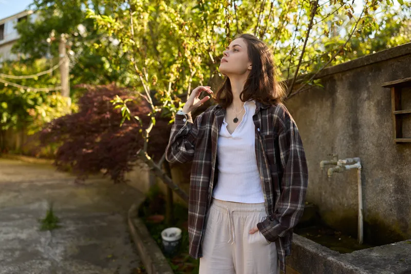 Casual woman outdoors, relaxed pose, sunlit moment in a lush garden, plaid shirt over white top, beige pants, carefree mood, gentle breeze, natural colors, warm light, serene atmosphere