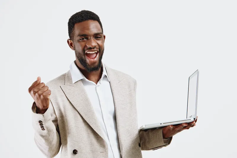 Confident happy young African American man wearing beige blazer and white shirt holding open laptop computer with cheerful expression, looking at camera, isolated on plain white background. People suc