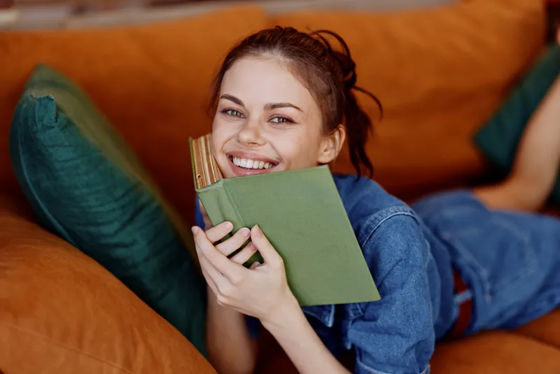 Счастливая женщина с улыбкой на лице наслаждается чтением книги, уютно устроившись на диване дома